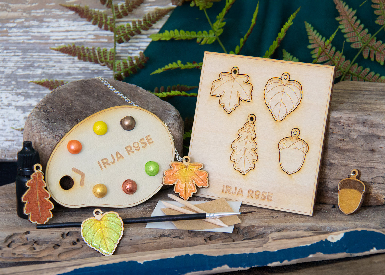 Wooden leaf-shaped charms and a paint palette with 'Irja Rose' branding on a wooden surface with ferns in the background.